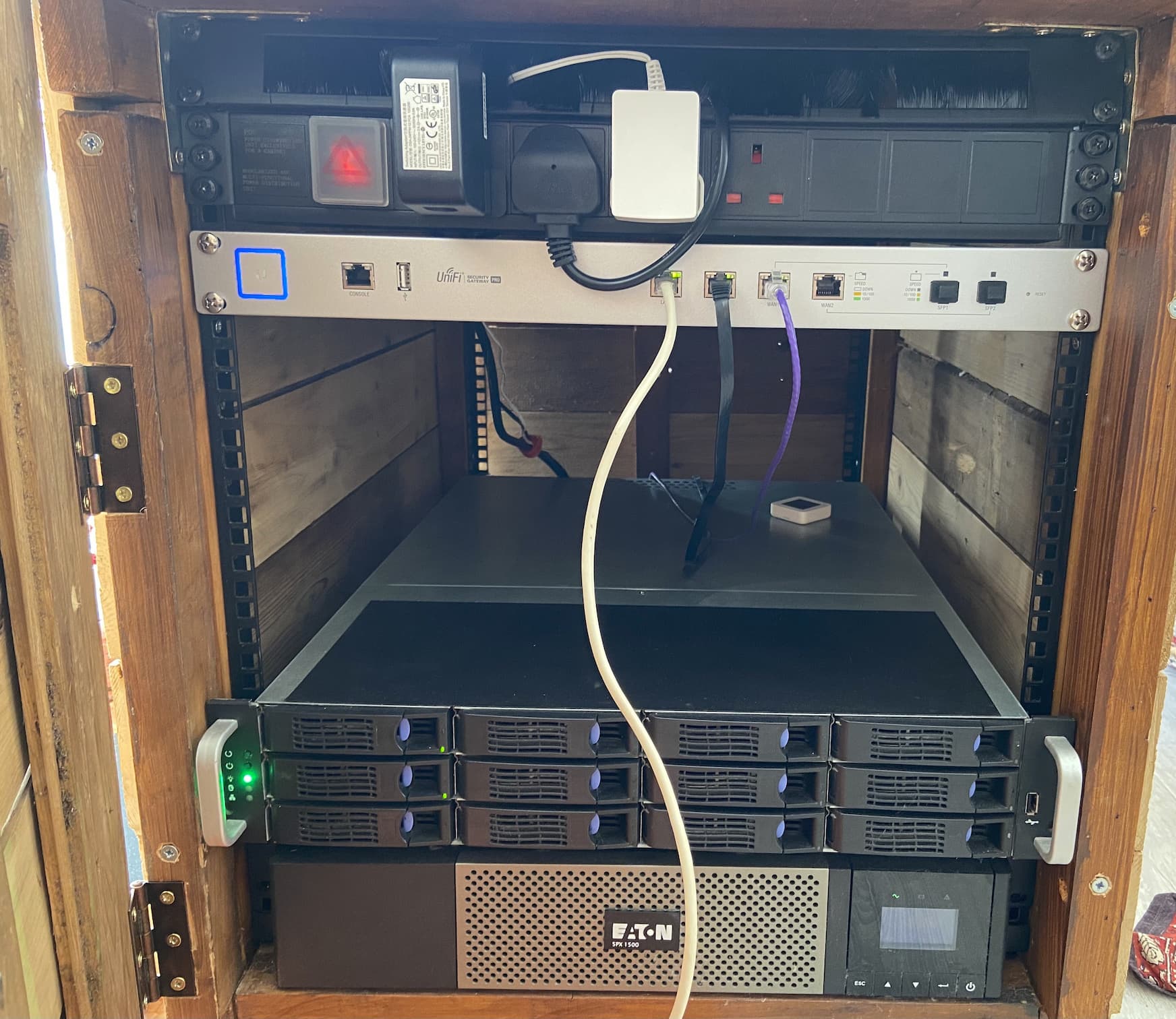A final picture of how things have progressed after twelve months, showing the original UPS and server mounted within, as well as a bluetooth temperature sensor, a PDU, a Unifi Security Gateway Pro and a bunch of cables