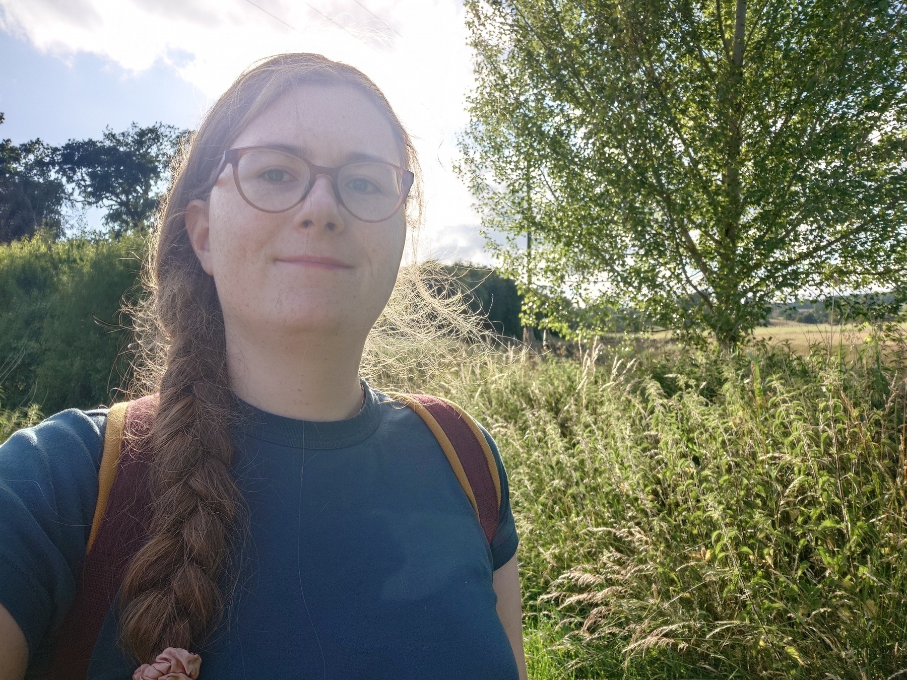 A selfie of Evie with a plain blue top next to a big tree in a field of overgrown.