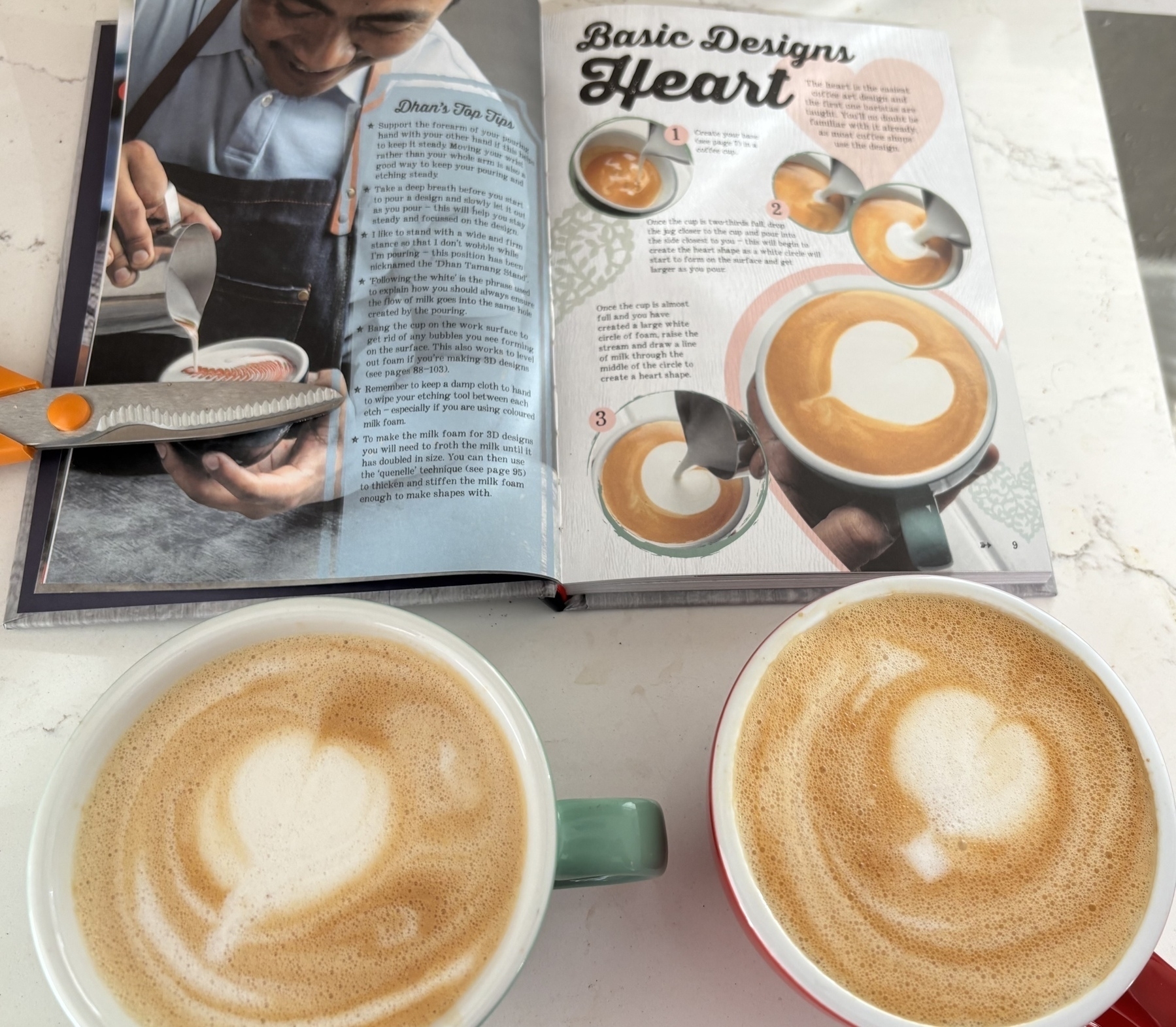 Two cups of coffee with heart latte art sit on a table next to an open book displaying latte art techniques.