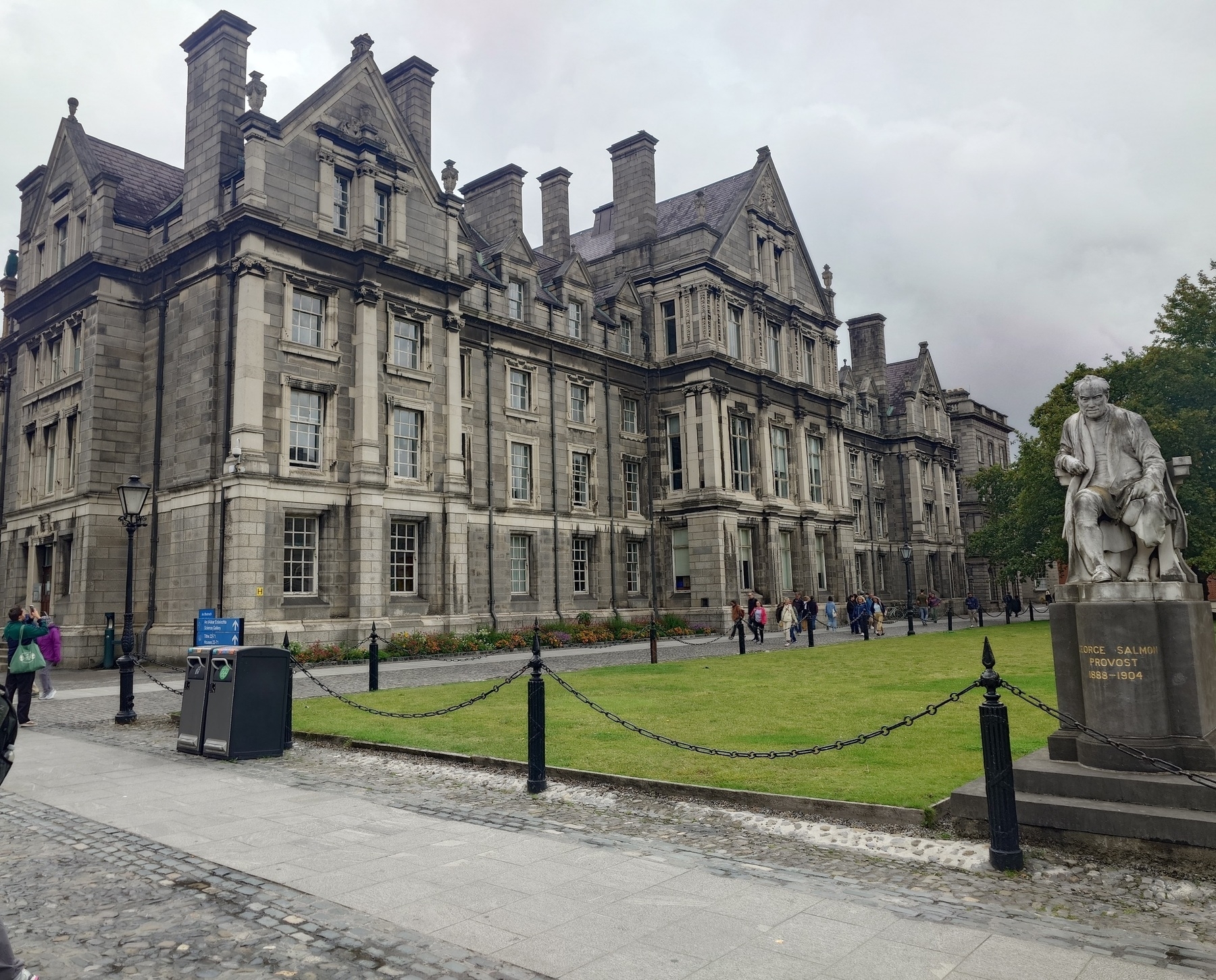 A historic stone building with a statue in front is surrounded by a grassy area and people walking by.