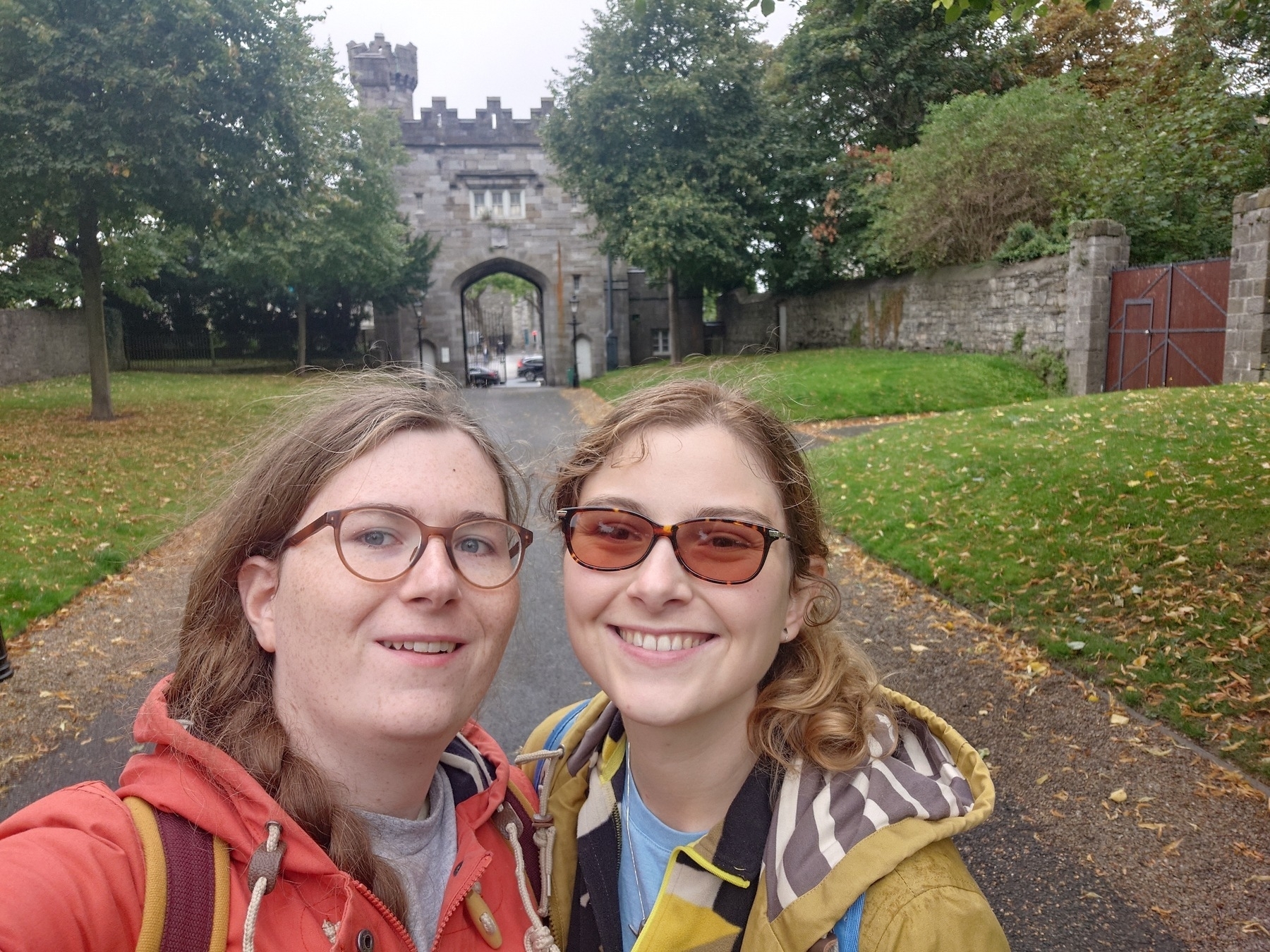 Two people wearing jackets pose for a selfie on a path leading to a stone archway surrounded by trees and grass.