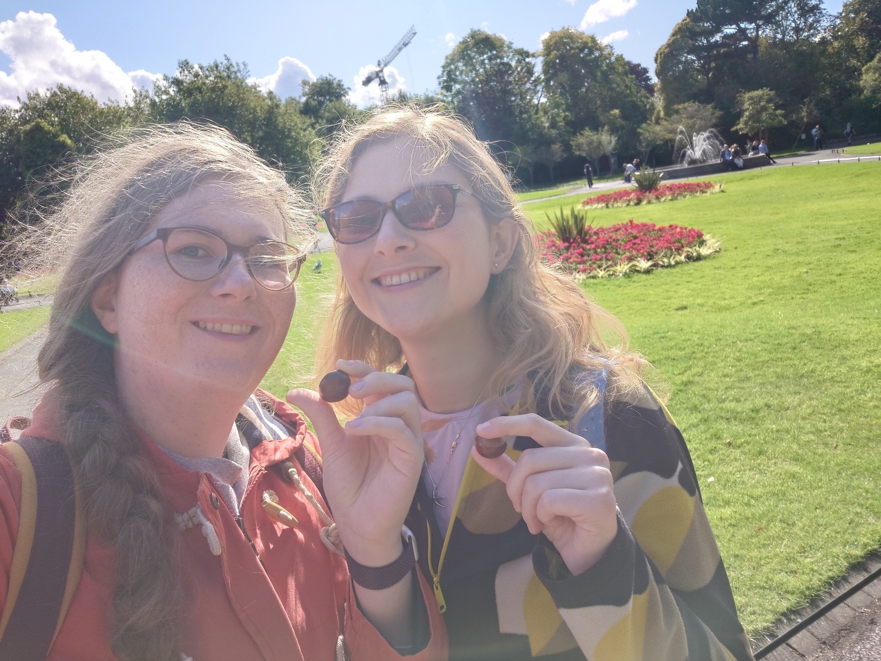 Two people smiling and holding small conkers in a sunny park with a well-maintained garden in the background.