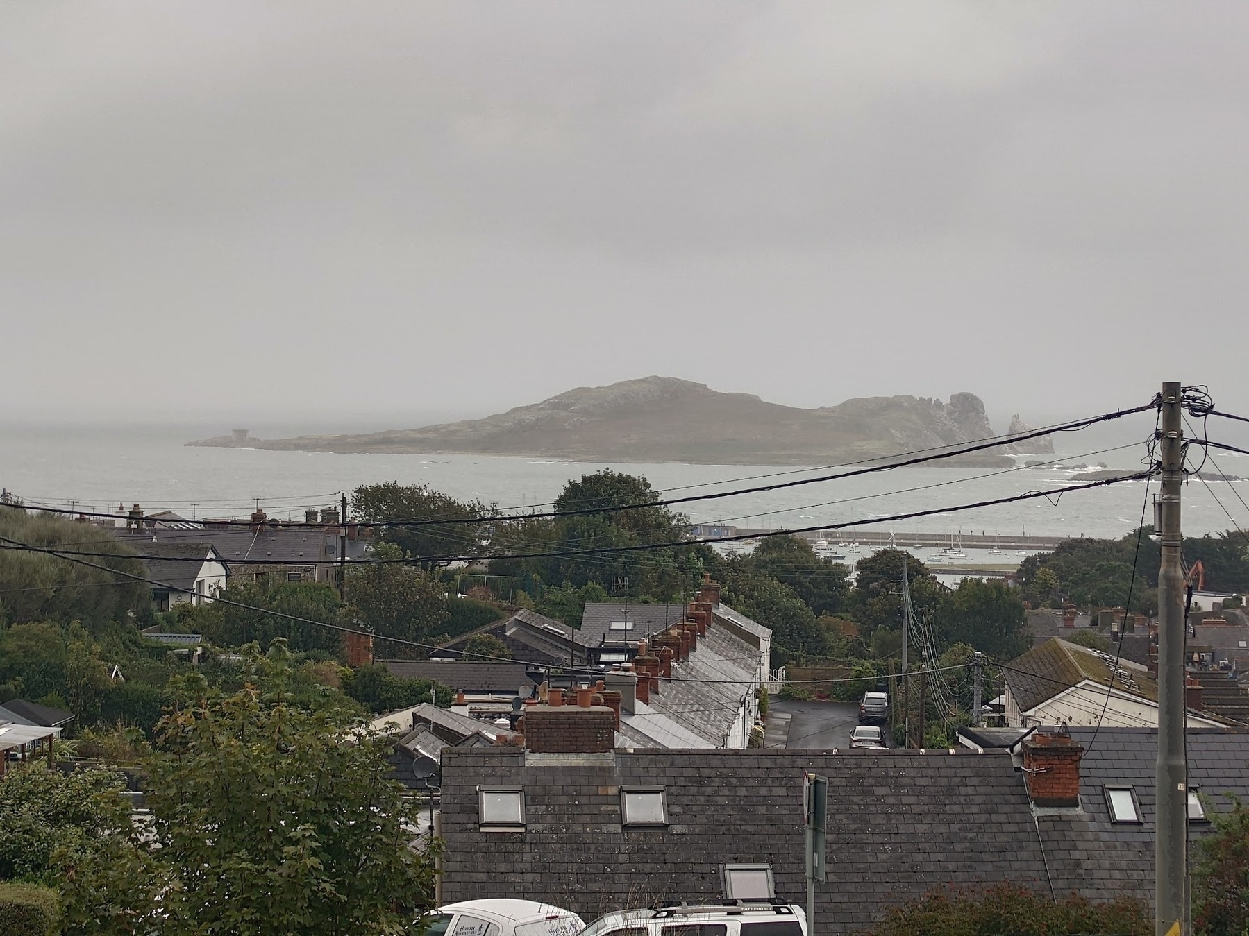 A coastal village overlooking the sea features rooftops, power lines, and a distant island under a cloudy sky.