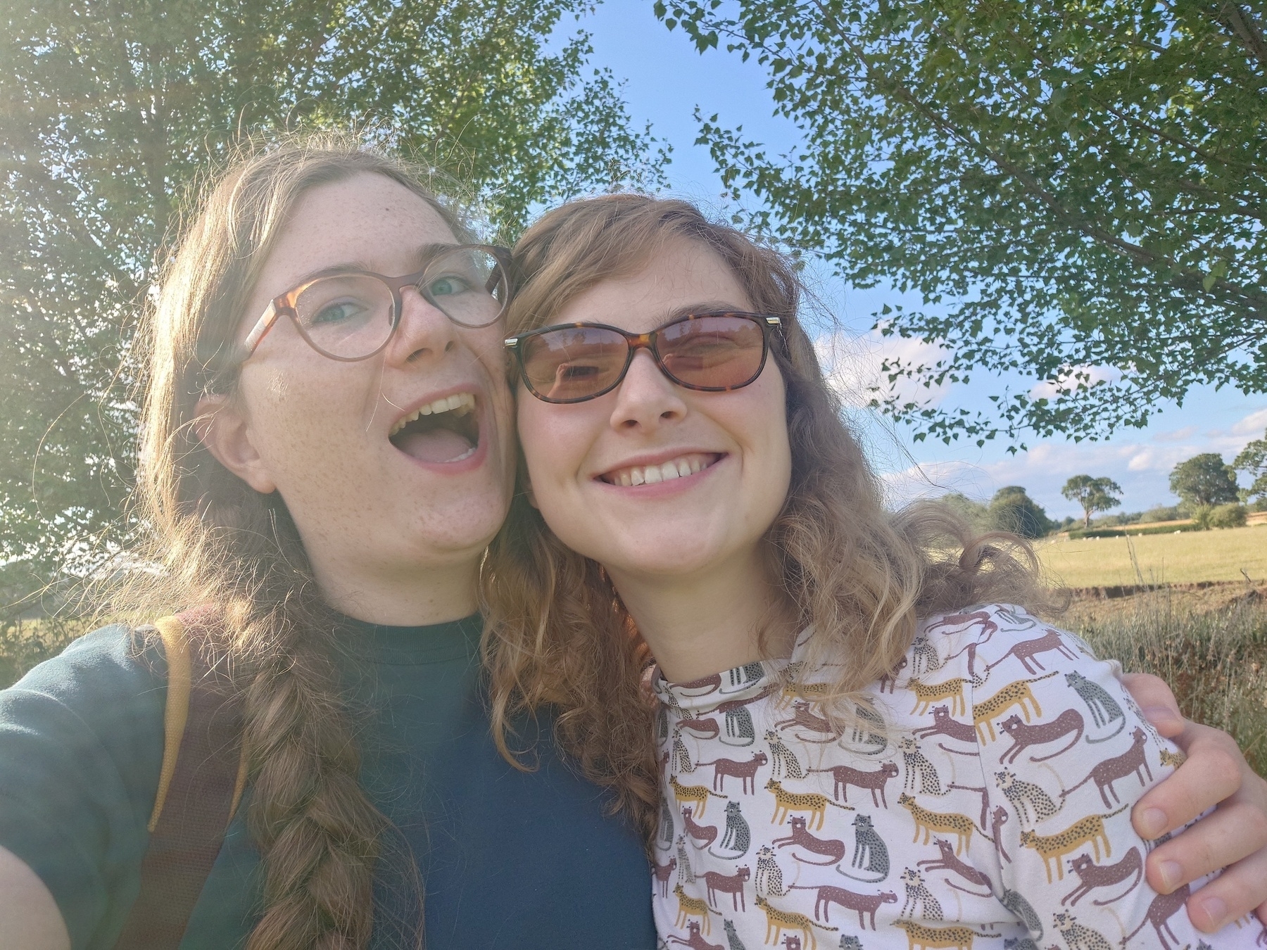 Two people are outdoors, smiling and posing for a selfie in a sunny field with trees in the background.