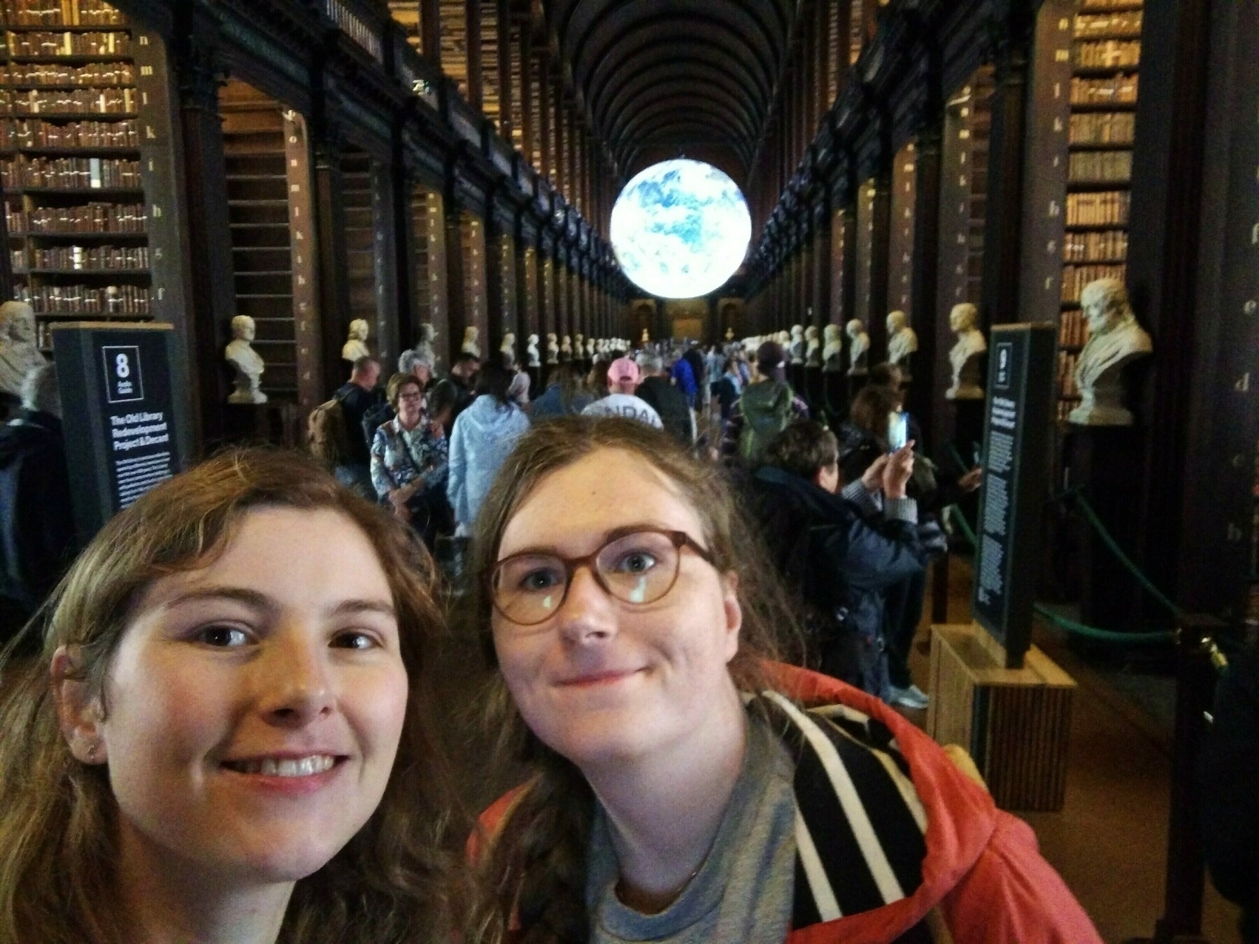 Two people are taking a selfie in a grand library lined with books and statues, with a large illuminated globe hanging in the background.