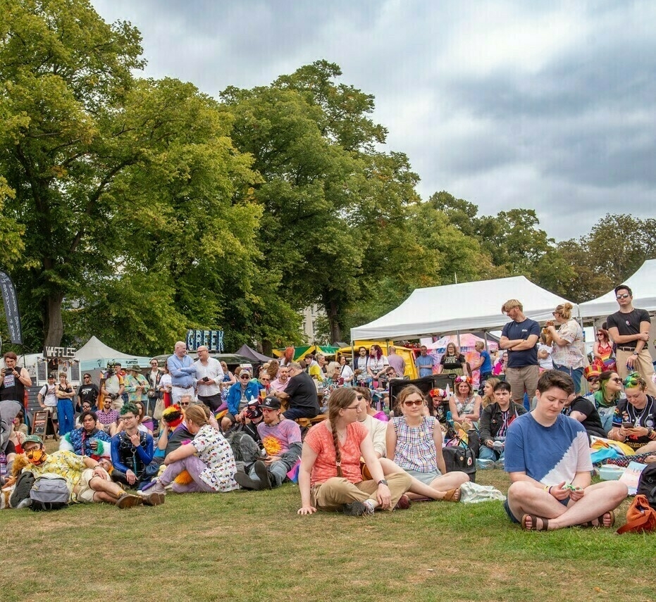 A large group of people is sitting on the grass enjoying an outdoor pride event with tents and trees in the background.