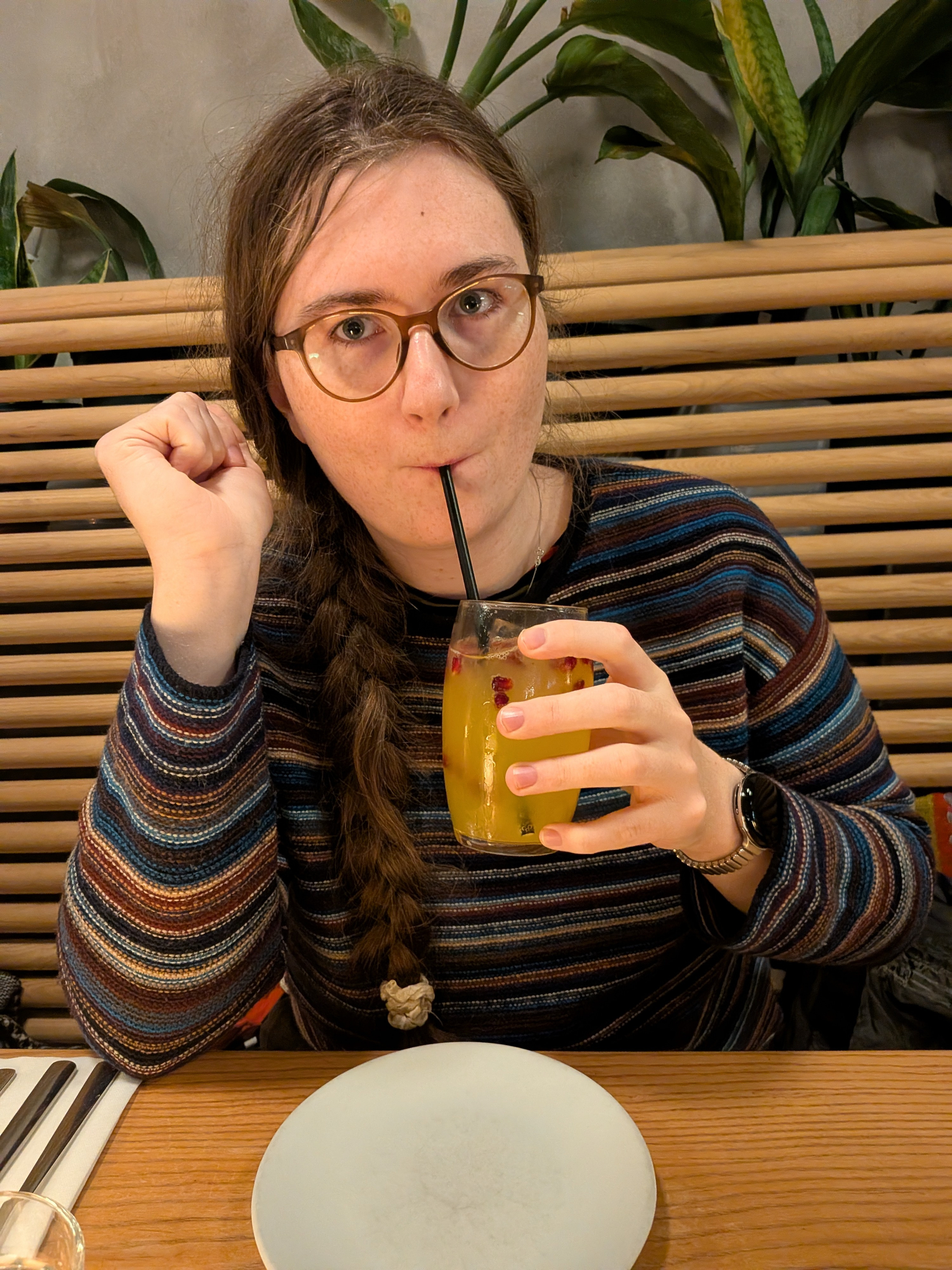 A person with glasses and a long braid sips a drink through a straw at a restaurant table.